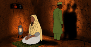 A woman sits on a prayer mat in a dimly lit, clay-walled room, illuminated by a small lantern. Her husband stands behind her, facing the wall, where his shadow splits into two overlapping silhouettes—one masculine, one feminine.